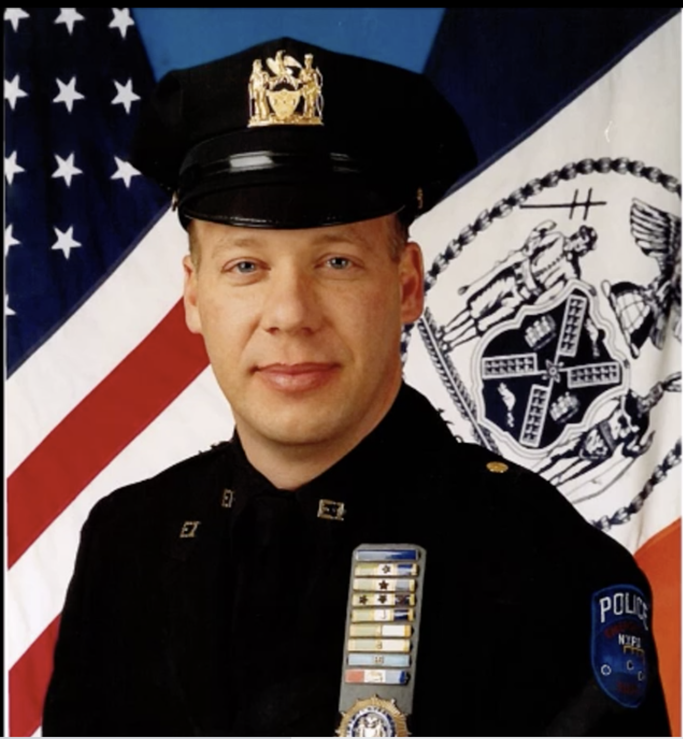 Police Detective Scott Strauss stands in front of an American Flag and the New York City Flag wearing a navy blue police uniform. The uniform includes a brimmed cap which features a gold medallion in the center, as well as a collared shirt with badge, nameplate, various colored honor and rank bars, and an ESU police patch on the left shoulder.pa