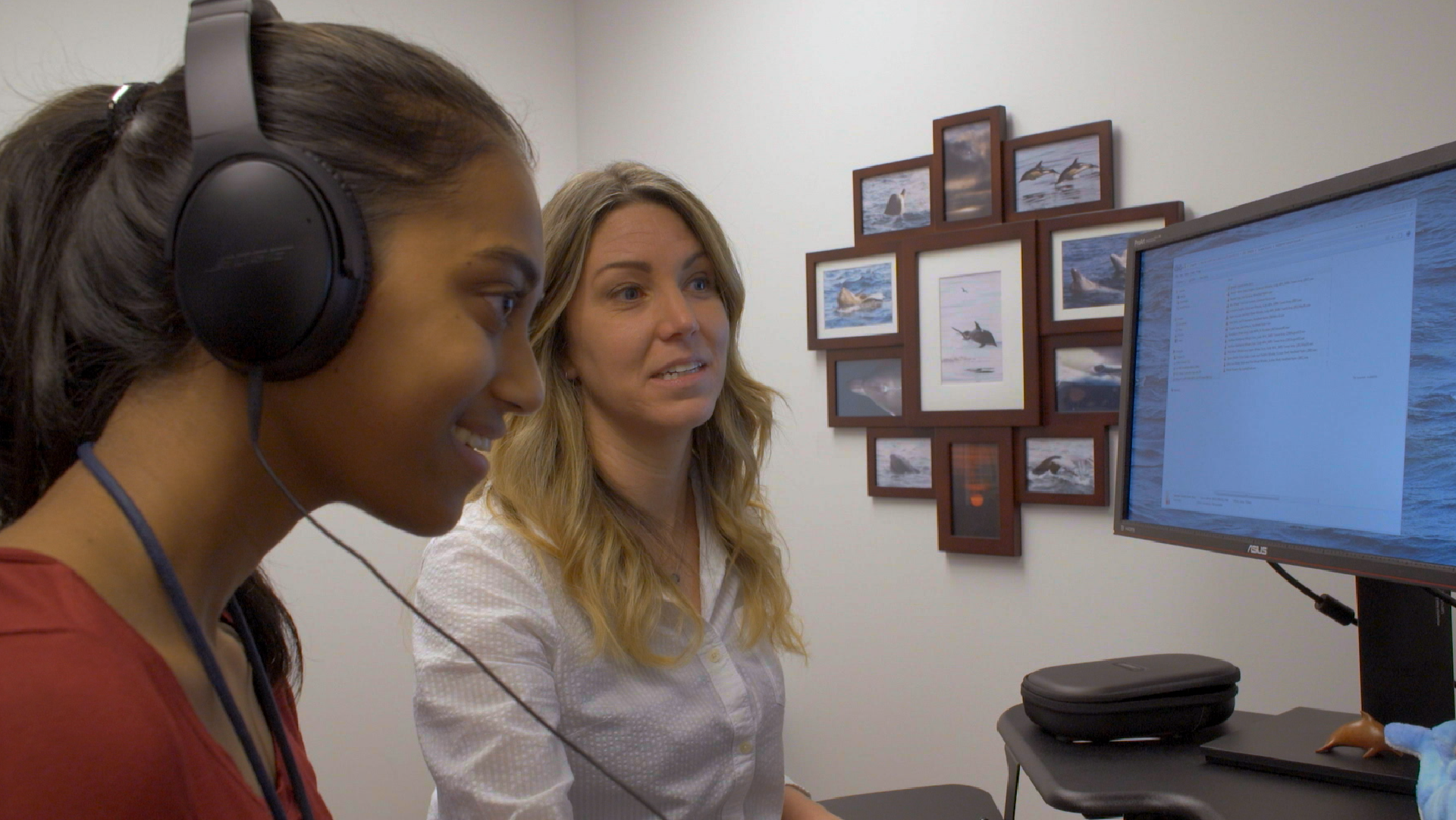 A woman sat besides a student wearing headphones and looking at a computer screen