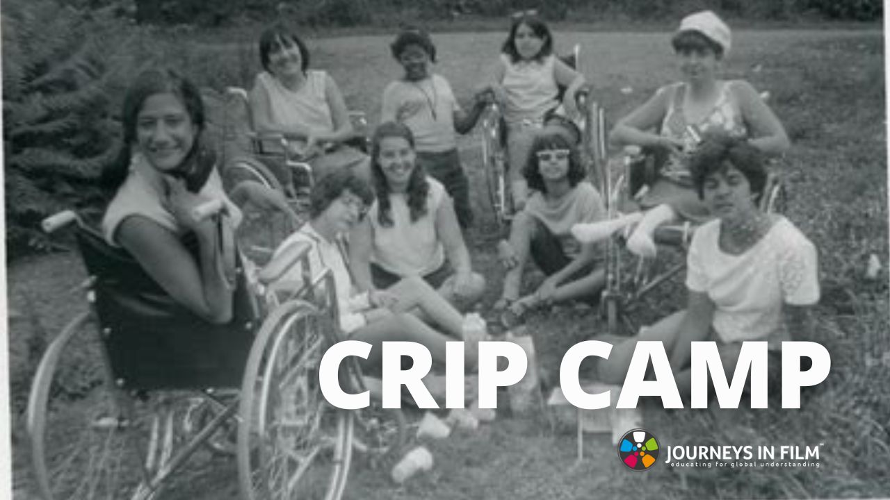 Vintage black and white photo from 1971 of a group of teen girls at Camp Jened, a summer camp in New York for teens with disabilities. Some are in wheelchairs and some sit on the ground. They are all smiling at the camera and look like they're having a great time. It's a fun, engaging photo. It says "Crip Camp" in bold white letters at the bottom, followed by the Journeys in Film logo.