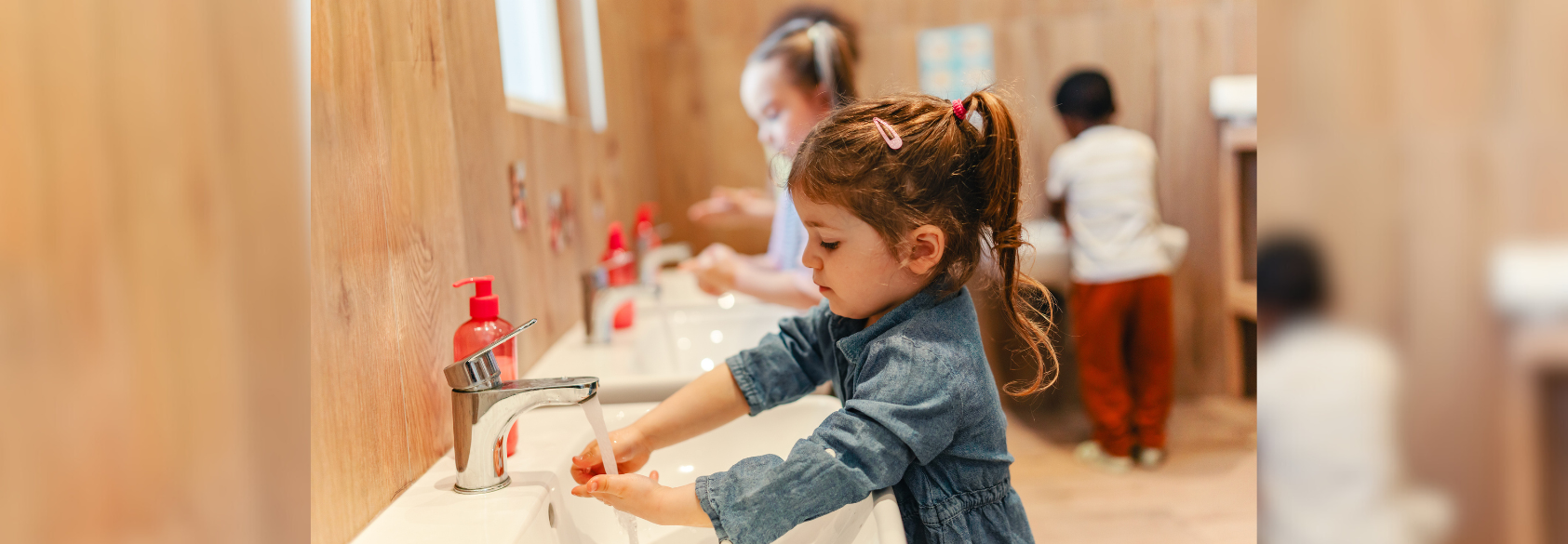 A young child washes their hands at a sink in a school bathroom, with other children using sinks in the background.