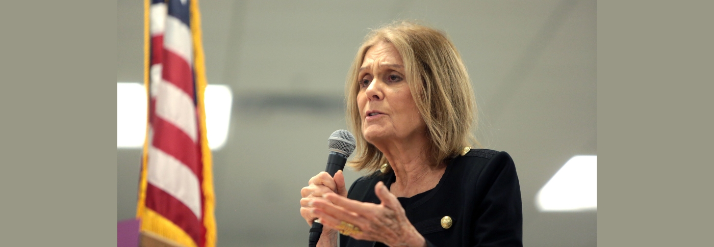 Gloria Steinem speaks into a handheld microphone at a public event, gesturing with one hand, a U.S. flag behind her.